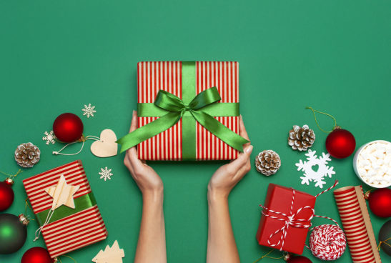 Hands holding present with Christmas gifts and ornaments around on a green background.