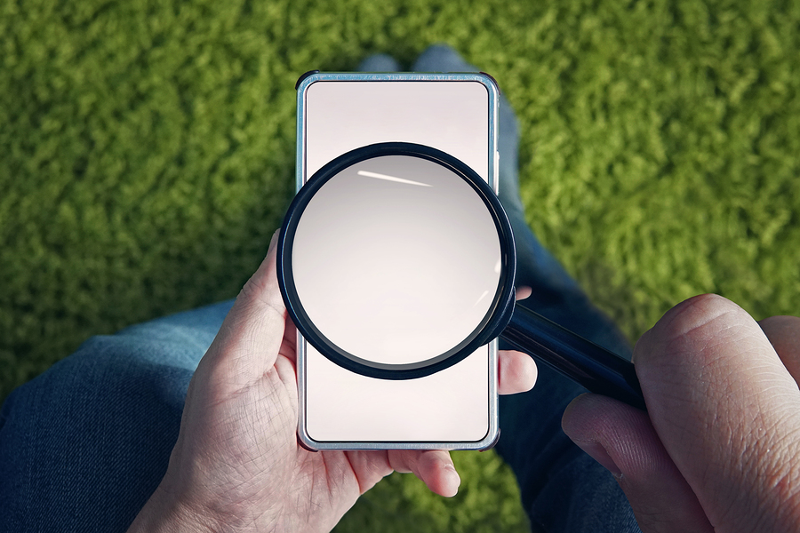 Man holding blank smartphone with magnifying glass above screen on grassy background.