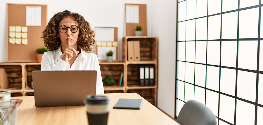 Image shows woman at work making the “be quiet” gesture.