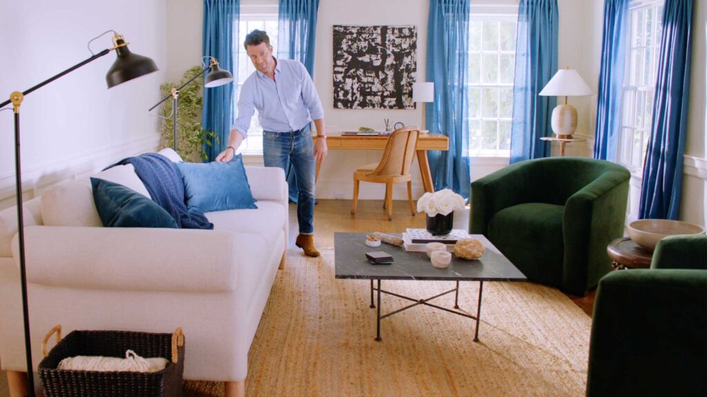 Nate Berkus fixing up a blue pillow on a couch in a living room.