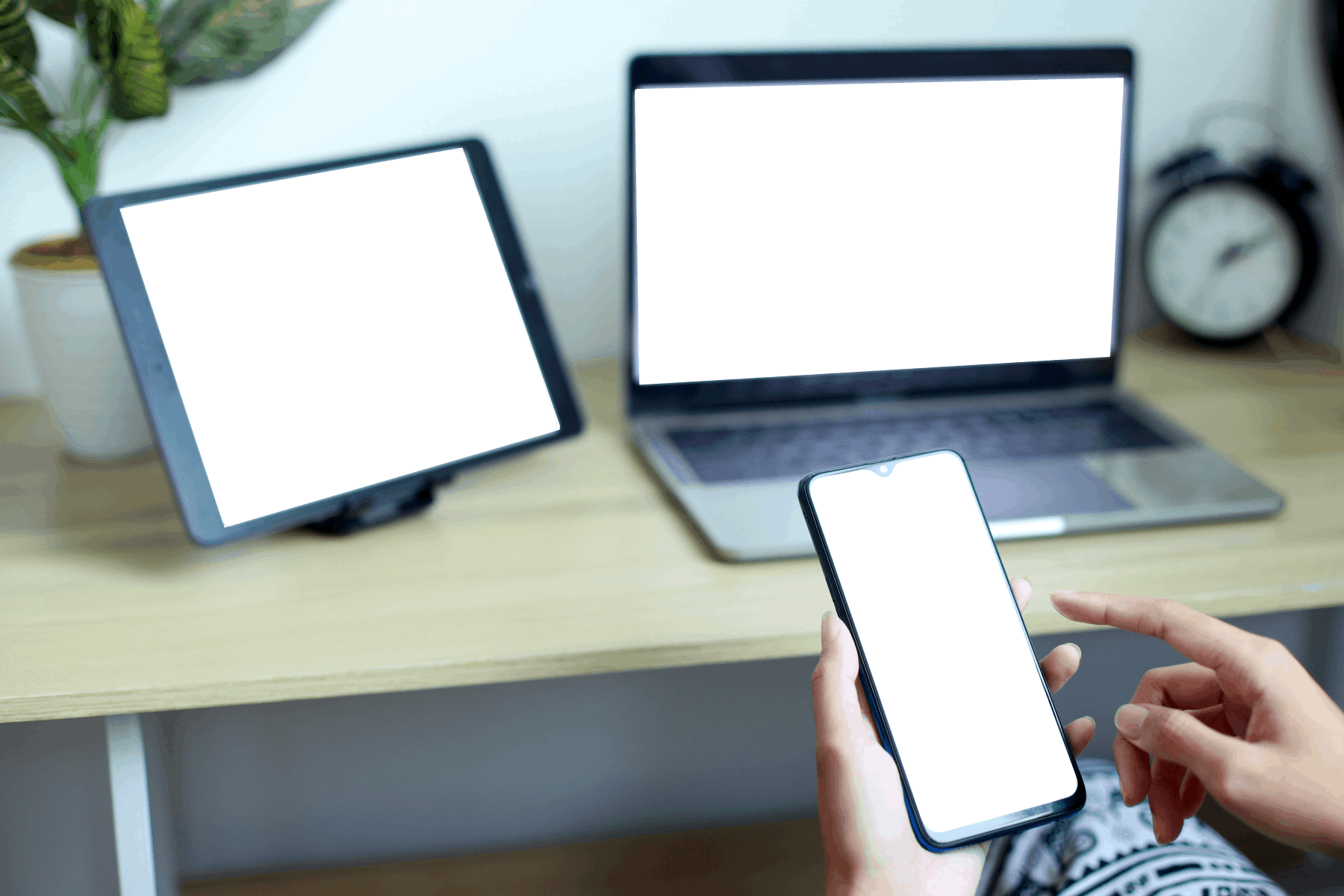 Hands holding a blank white screen mobile phone with laptop computer and tablet on the desk